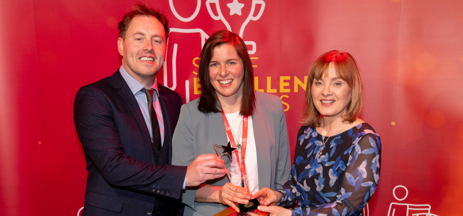 two women and one man, smartly dressed, in front of a red background